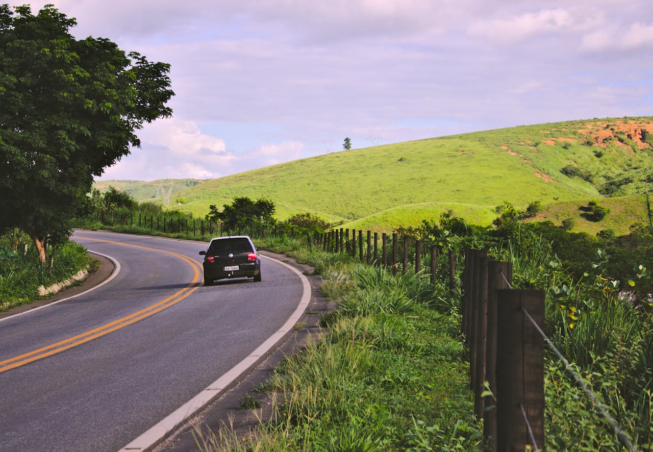 A car travels on a winding rural road in lush Brazilian countryside under a blue sky.